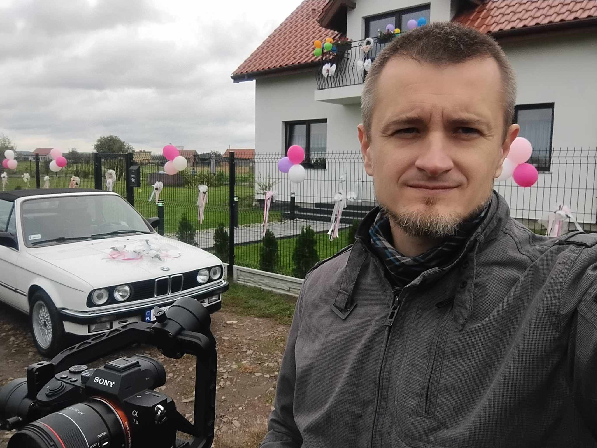 Uśmiechnięty mężczyzna z brodą robiący selfie z kamerą Sony na stabilizatorze, w tle białe BMW kabriolet udekorowane na ślub, dom z balkonem i balonami.