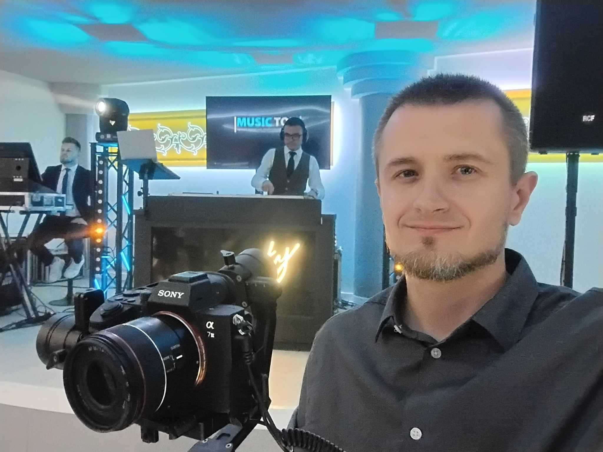 Kamerzysta z uśmiechem prezentuje kamerę Sony Alpha 7 III na statywie, w tle DJ na stanowisku z napisem MUSIC TO