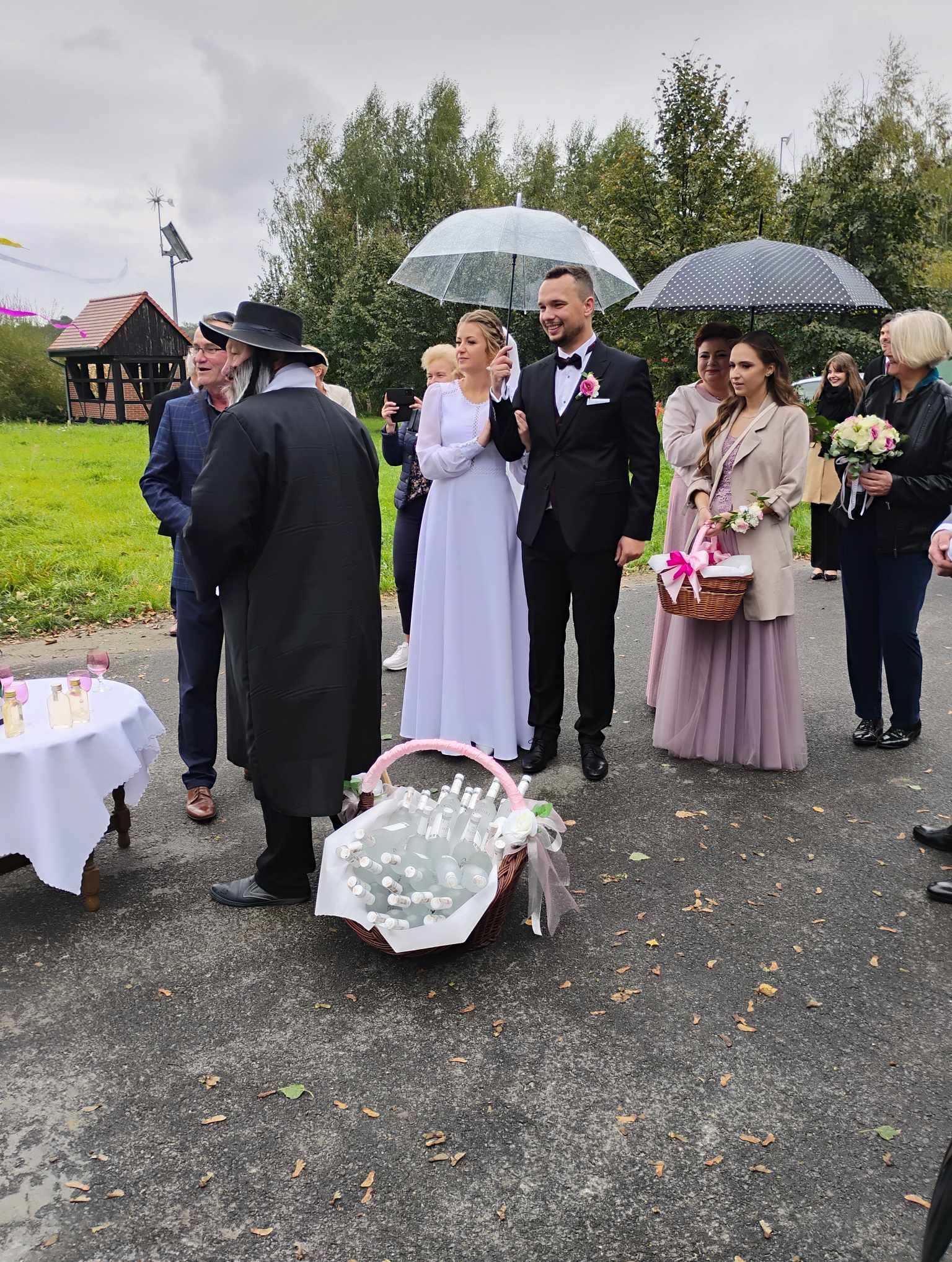 Para młoda pod przezroczystymi parasolami wychodzi z ceremonii, orszak weselny z koszykami i bukietami, butelki z alkoholem na stole, pochmurny dzień.