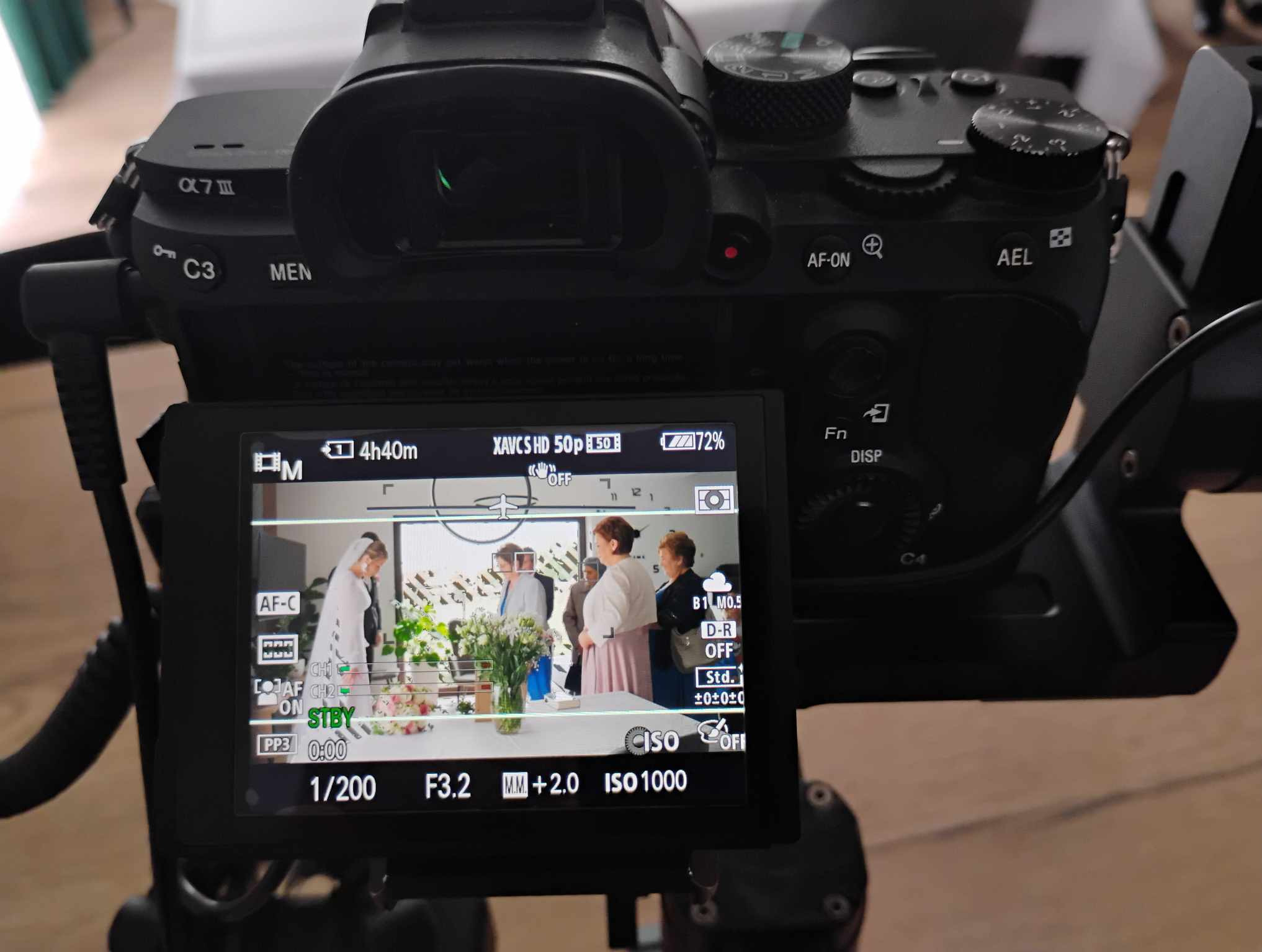 Ekran kamery Sony Alpha 7 III z widokiem na nagrywaną scenę ślubną: panna młoda w białej sukni, goście, kwiaty na stole. Ustawienia kamery: 1/200, F3.2, ISO 1000.