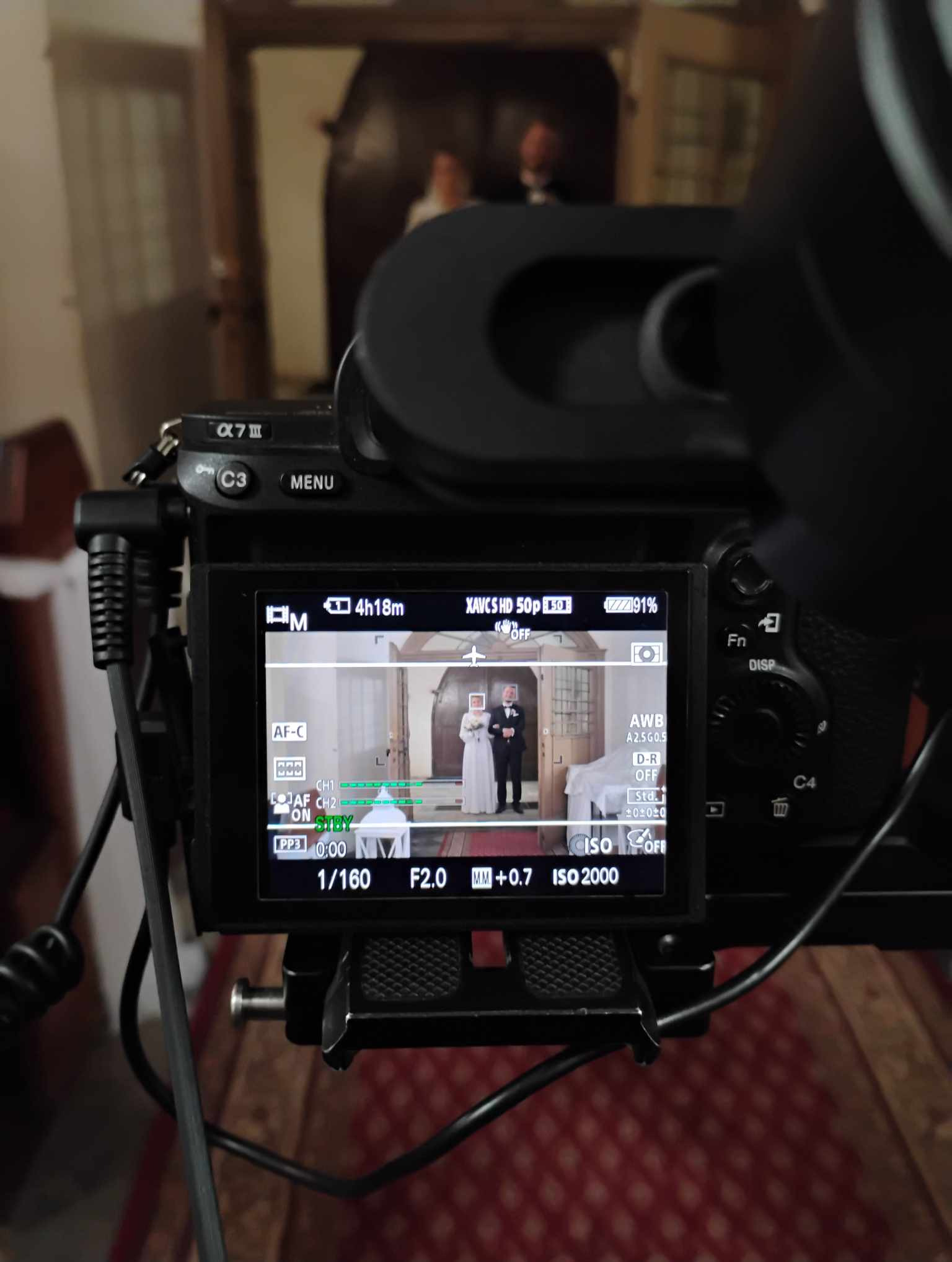 Ekran kamery Sony Alpha 7 III z widokiem na parę młodą w eleganckim wnętrzu, ustawienia ekspozycji: 1/160s, f/2.0, ISO 2000, na pierwszym planie fragment kamery i statywu.