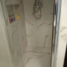 Kabina prysznicowa z marmurowymi płytkami i odpływem liniowym w podłodze, widoczne kosmetyki w półce ściennej.