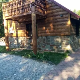 Drewniany dom z balkonem, elewacja z bali i kamienną podmurówką, widoczne oświetlenie zewnętrzne i chodnik z kostki.