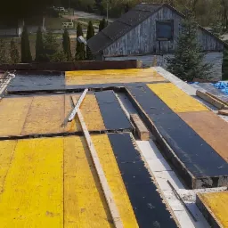 Widok z góry na szalunek stropu z żółtymi i czarnymi płytami, zbrojenie stalowe i poziomica, w tle budynek gospodarczy i drzewa.