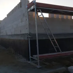 Budowa ściany z bloczków silikatowych na fundamencie, widoczne rusztowanie, palety z bloczkami w tle, wieczorne oświetlenie.