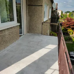 Wyłożony jasnymi płytkami balkon z brązową barierką, widok na zieleń i domy w tle.