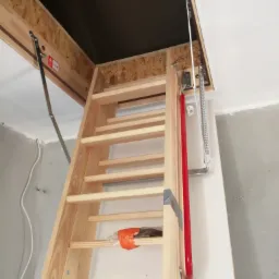 Drewniane schody strychowe składane, widoczne mechanizmy sprężynowe i czerwony amortyzator, zamontowane w suficie z płyt OSB.