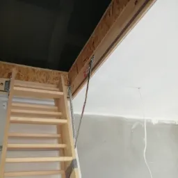 Drewniane schody strychowe rozkładane, widoczny mechanizm sprężynowy, sufit z płyt karton-gips, ściana w trakcie szpachlowania.