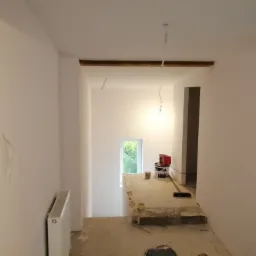 Wnętrze w trakcie remontu: pomalowane ściany na biało, widoczne puszki z farbą, grzejnik, fragment drewnianej konstrukcji pod sufitem i wystające przewody.