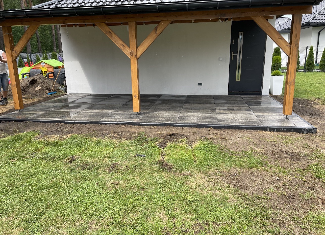 Ułożona, szara kostka brukowa pod zadaszeniem domu, widok z poziomu trawnika. Widoczne drewniane podpory zadaszenia i fragment elewacji budynku.