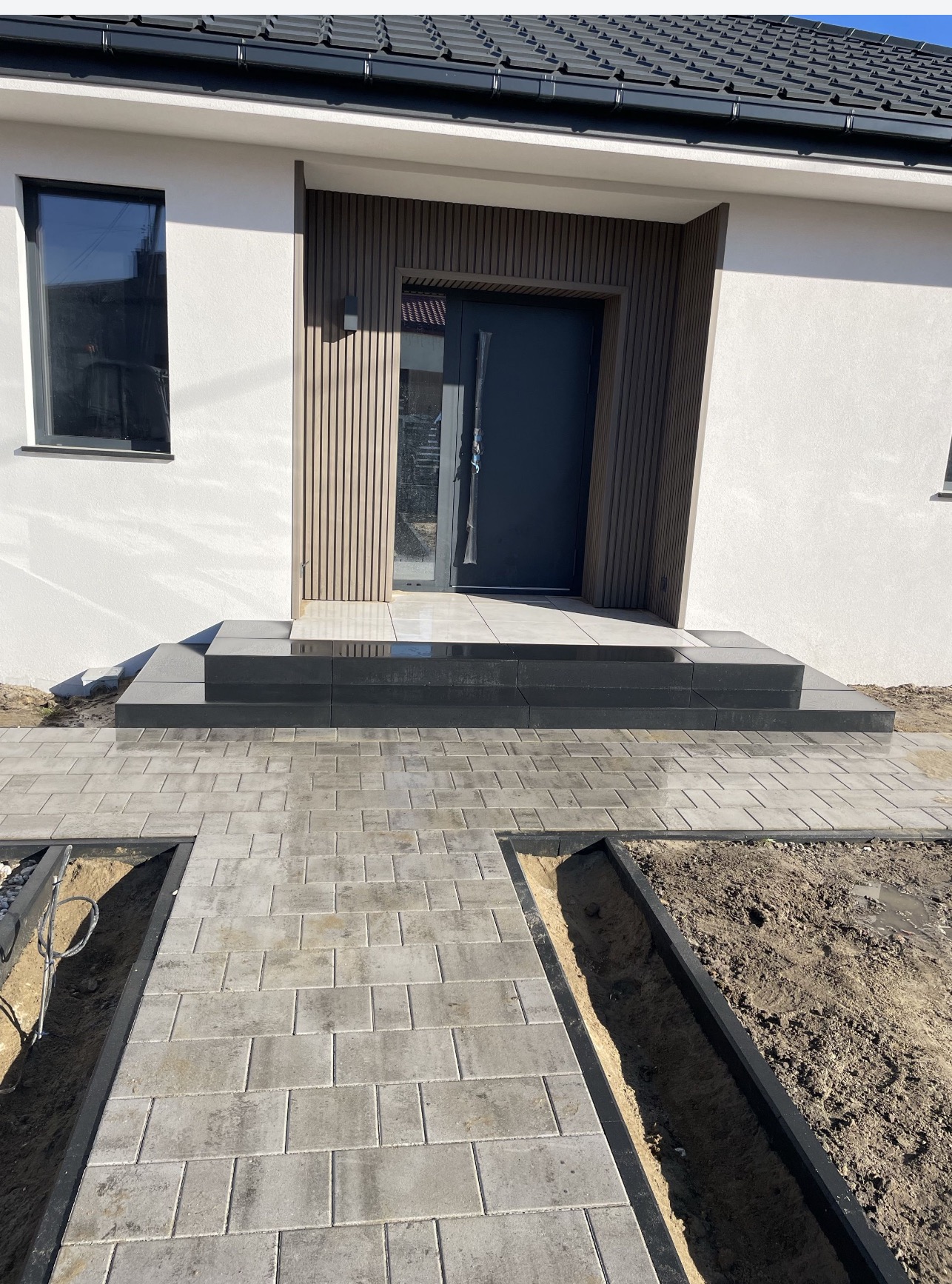 Nowoczesne wejście do domu z szarej kostki brukowej, czarne obramowanie rabat, minimalistyczne schody i drzwi wejściowe z drewnianą okładziną.