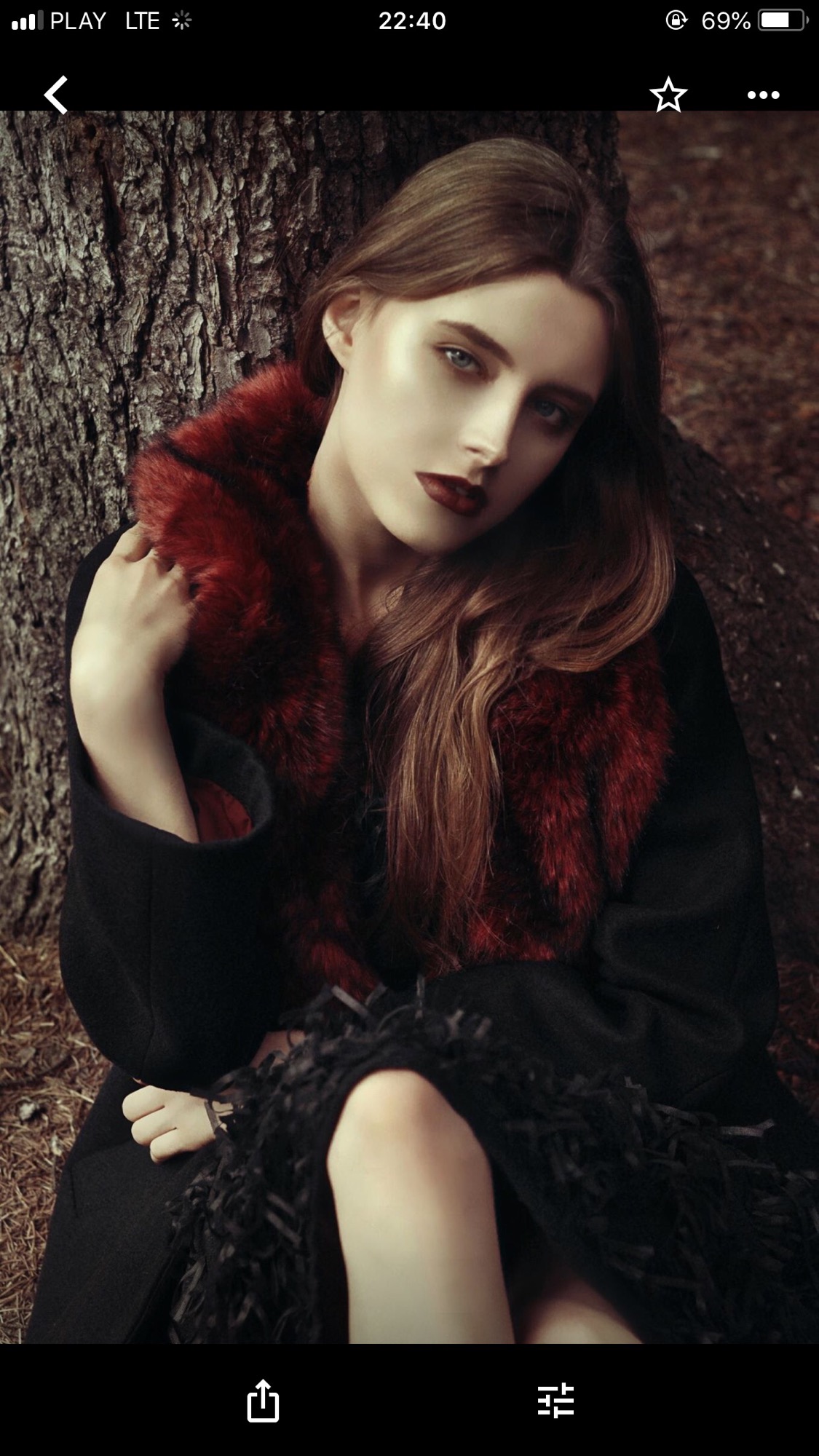 Portret kobiety z bladą cerą i ciemną pomadką, w czarnym płaszczu z bordowym futrzanym kołnierzem, siedzącej pod drzewem.