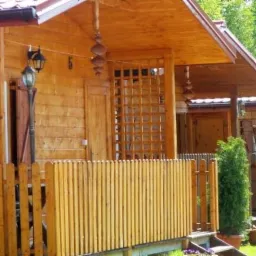 Drewniany domek letniskowy z numerem 5 nad drzwiami, ozdobiony drewnianą kratą i wiszącą dekoracją, otoczony niskim płotkiem i zielenią.