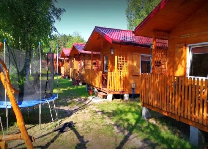 Rząd drewnianych domków letniskowych z czerwonymi dachami i drewnianymi werandami, z trampoliną i huśtawką na pierwszym planie, otoczonych zielenią.