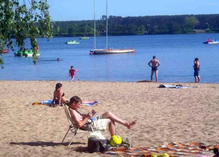 Plaża nad jeziorem w Sielpi Wielkiej: wypoczywający ludzie, łódki na wodzie, las w tle.
