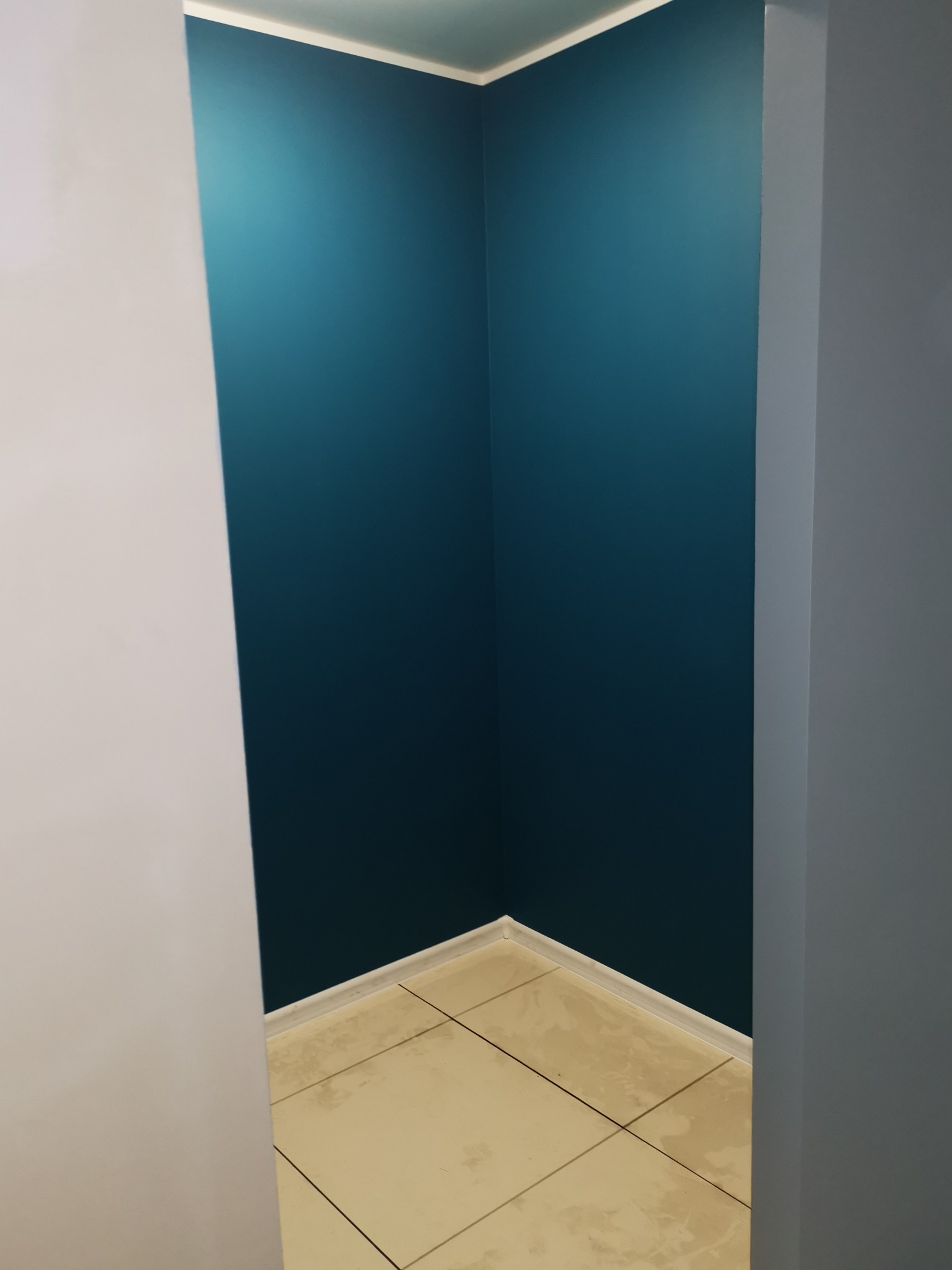 Narożnik pomalowany na intensywny, ciemny odcień niebieskiego w mieszkaniu w Świerznie, widoczne jasne płytki podłogowe i biała listwa przypodłogowa, kontrastujące z szarymi elementami...