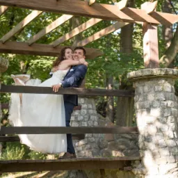 Uśmiechnięta panna młoda w białej sukni, niesiona przez pana młodego na drewnianym mostku z kamiennymi filarami w parku, wśród zieleni drzew.