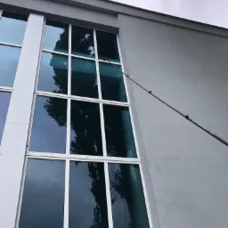 Wysokie okna biurowca myte z zewnątrz za pomocą teleskopowego kija z szczotką, widoczne smugi wody na szybach.