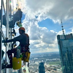 Na Linie Michał Ciechanowski - Pracownicy wysokościowi myjący okna drapacza chmur w słoneczny dzień. Widok na panoramę miasta z góry. Żółte wiadro i sprzęt alpinistyczny.