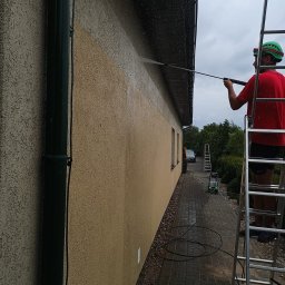 Na Linie Michał Ciechanowski - Pracownik na drabinie czyści elewację domu myjką ciśnieniową. Widoczny fragment elewacji przed i po czyszczeniu. W tle zaparkowany samochód i dodatkowa drabina.