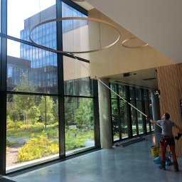 Na Linie Michał Ciechanowski - Pracownik w trakcie mycia wysokich okien w biurowcu, używając specjalistycznego sprzętu. Widoczne duże okna z widokiem na zieleń, drabina i wiadro z akcesoriami.