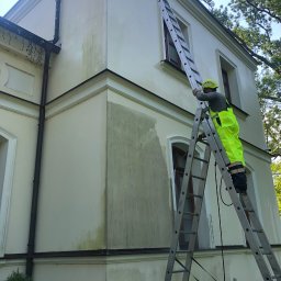 Na Linie Michał Ciechanowski - Pracownik w żółtym kombinezonie na drabinie, myjący fragment elewacji budynku. Widoczne zabrudzenia na ścianie, okna z brązowymi ramami i fragmenty zieleni.