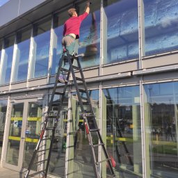 Na Linie Michał Ciechanowski - Pracownik na rozstawionej drabinie myje okna budynku biurowego. Widoczne odbicia w szybach i zabezpieczenia na oknach. Słoneczny dzień.