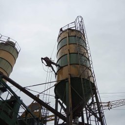 Na Linie Michał Ciechanowski - Pracownik na linie, zabezpieczony uprzężą, wykonuje prace na wysokości na silosie cementowym. Widoczne drabiny, liny i konstrukcja stalowa. Pochmurne niebo w tle.