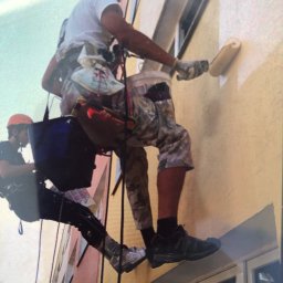 Na Linie Michał Ciechanowski - Dwóch pracowników na linach maluje elewację budynku wałkiem. Jeden maluje, drugi asekuruje. Widoczne okna i fragmenty innych budynków w tle.