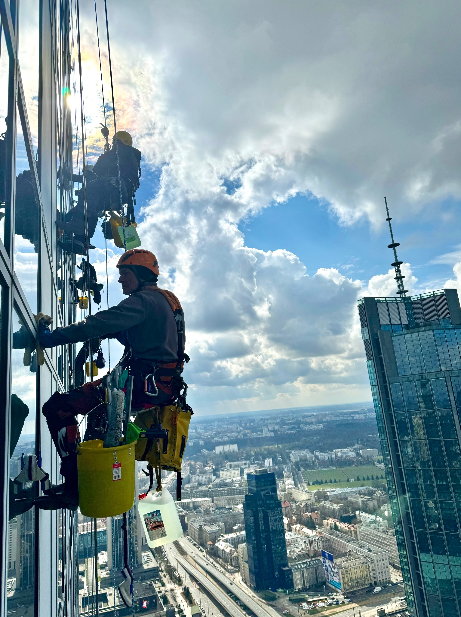 Pracownicy wysokościowi myjący okna drapacza chmur w słoneczny dzień. Widok na panoramę miasta z góry. Żółte wiadro i sprzęt alpinistyczny.