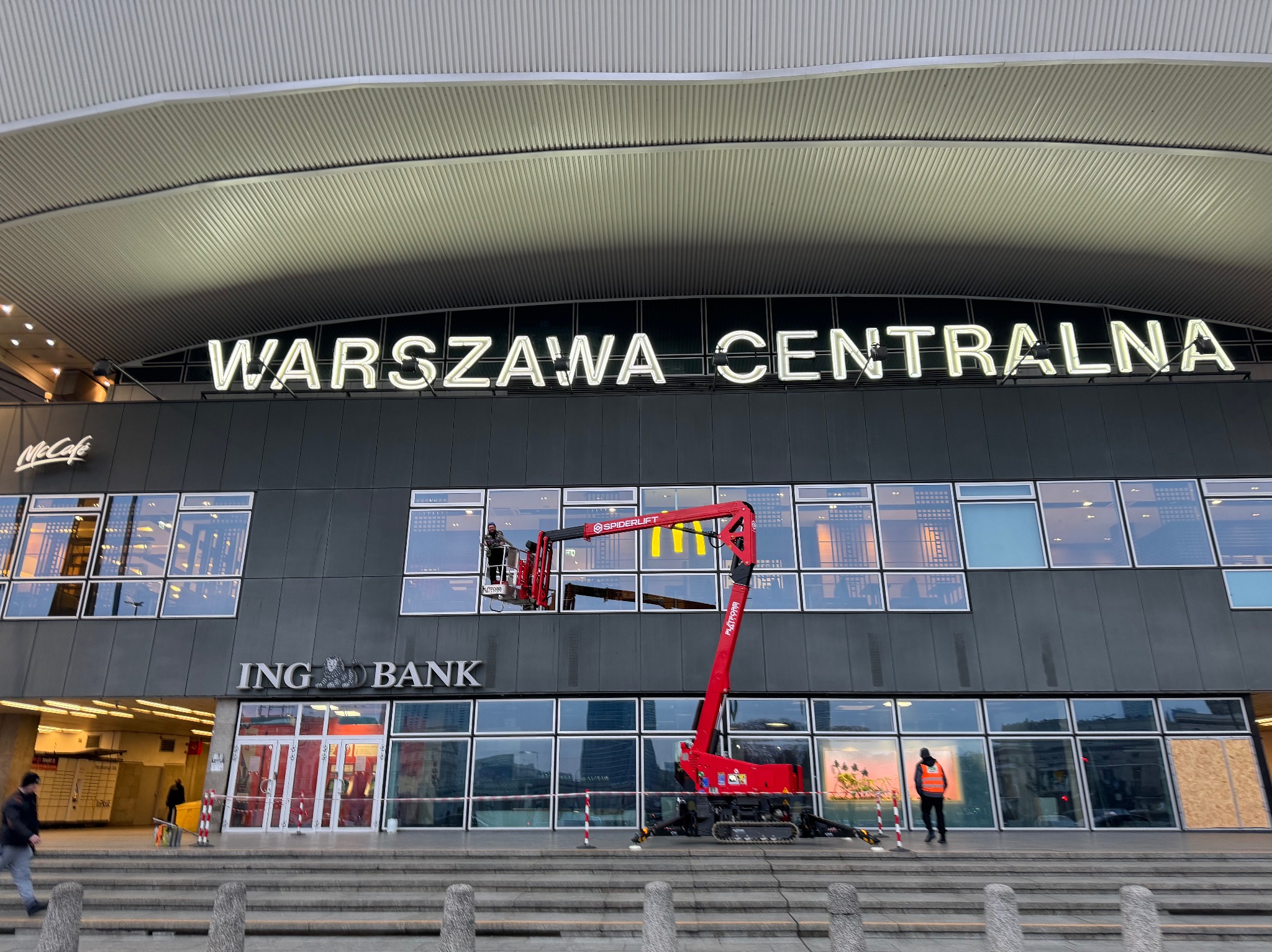 Czerwony podnośnik koszowy przed budynkiem Warszawa Centralna, pracownik w koszu. Widoczne okna, szyld ING Banku i McDonald's. Osoba w kamizelce na chodniku.