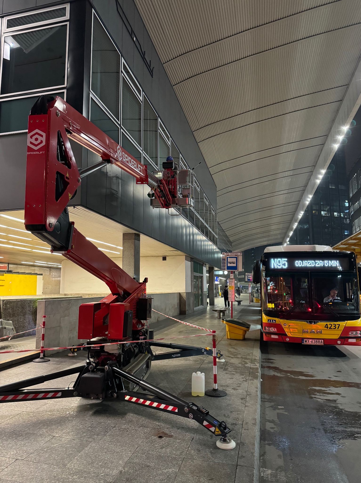 Czerwony podnośnik koszowy Spiderlift przy budynku dworca, obok autobus N95. Prace na wysokościach w przestrzeni miejskiej, widoczne zabezpieczenia.