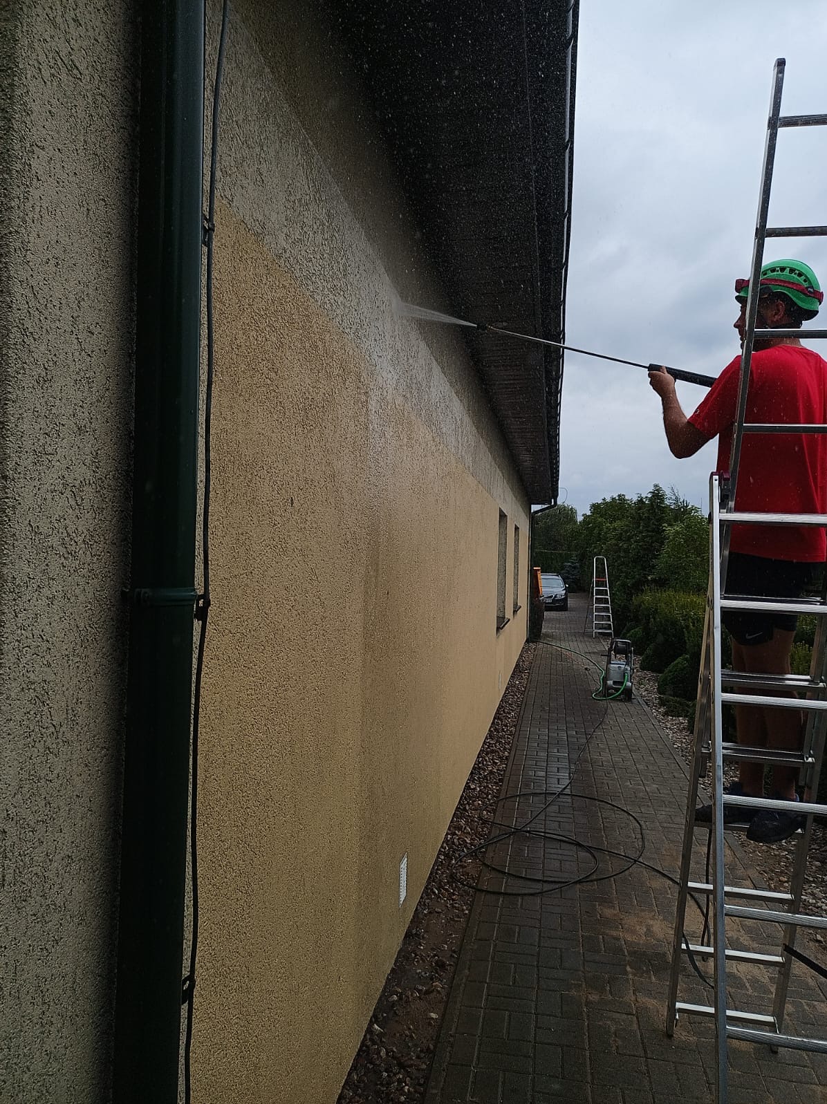 Pracownik na drabinie czyści elewację domu myjką ciśnieniową. Widoczny fragment elewacji przed i po czyszczeniu. W tle zaparkowany samochód i dodatkowa drabina.