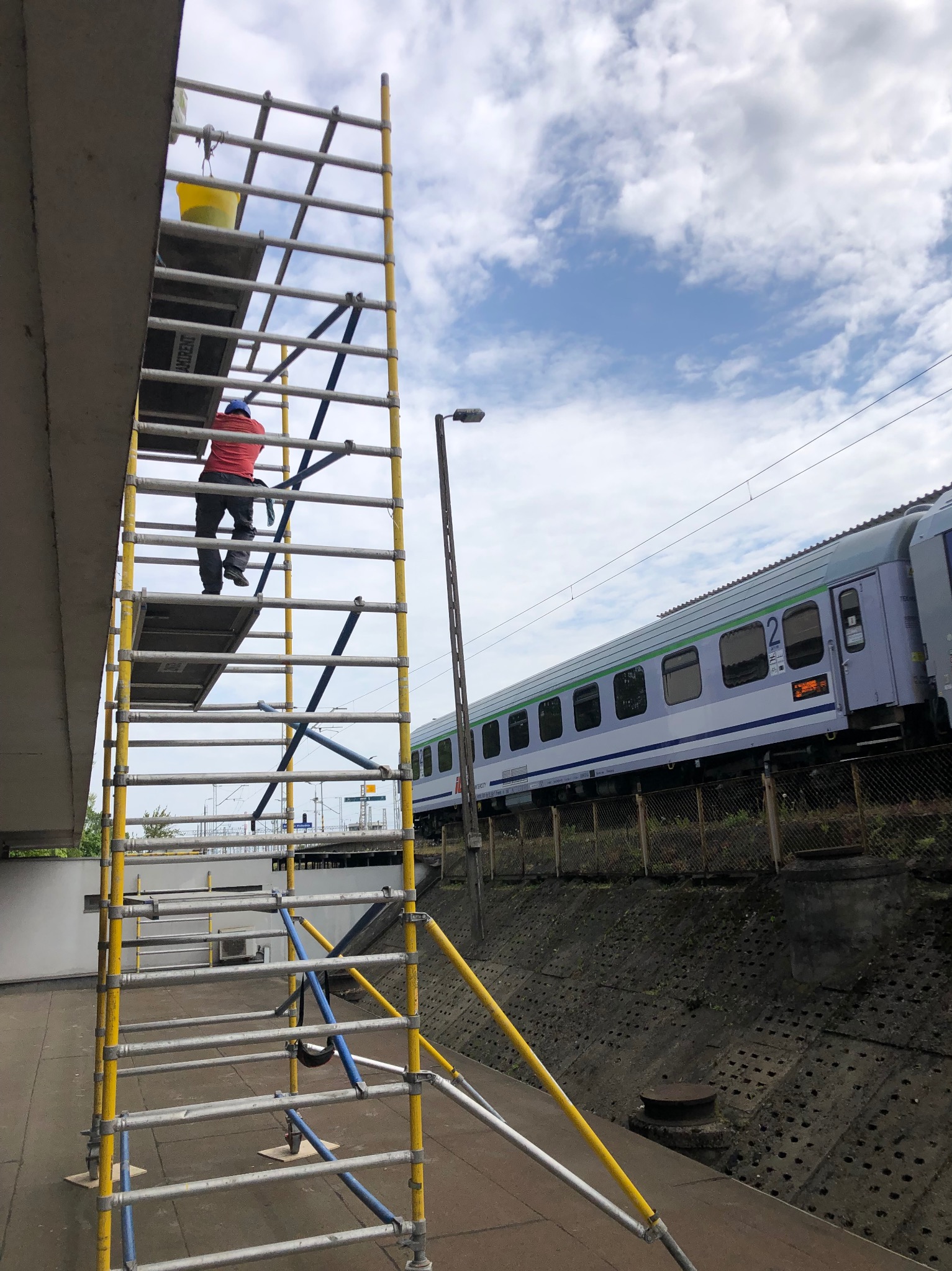 Pracownik na rusztowaniu przy budynku dworca, prawdopodobnie wykonuje prace konserwacyjne lub czyszczenie. W tle widoczny pociąg.