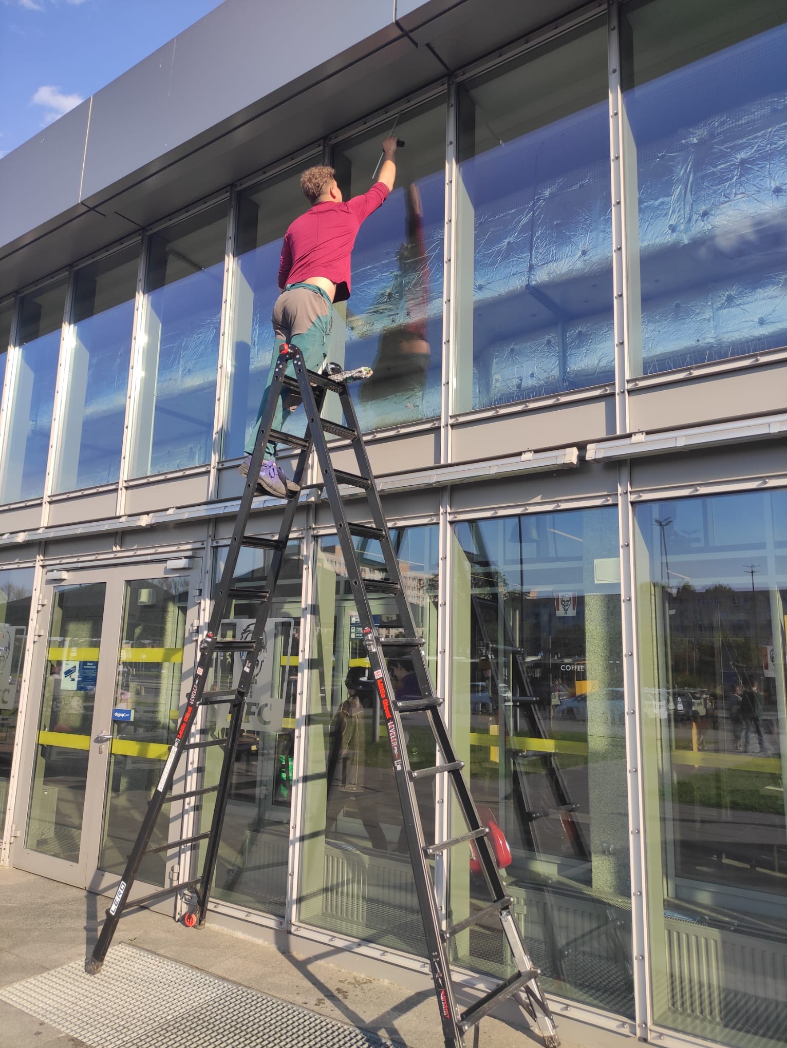 Pracownik na rozstawionej drabinie myje okna budynku biurowego. Widoczne odbicia w szybach i zabezpieczenia na oknach. Słoneczny dzień.