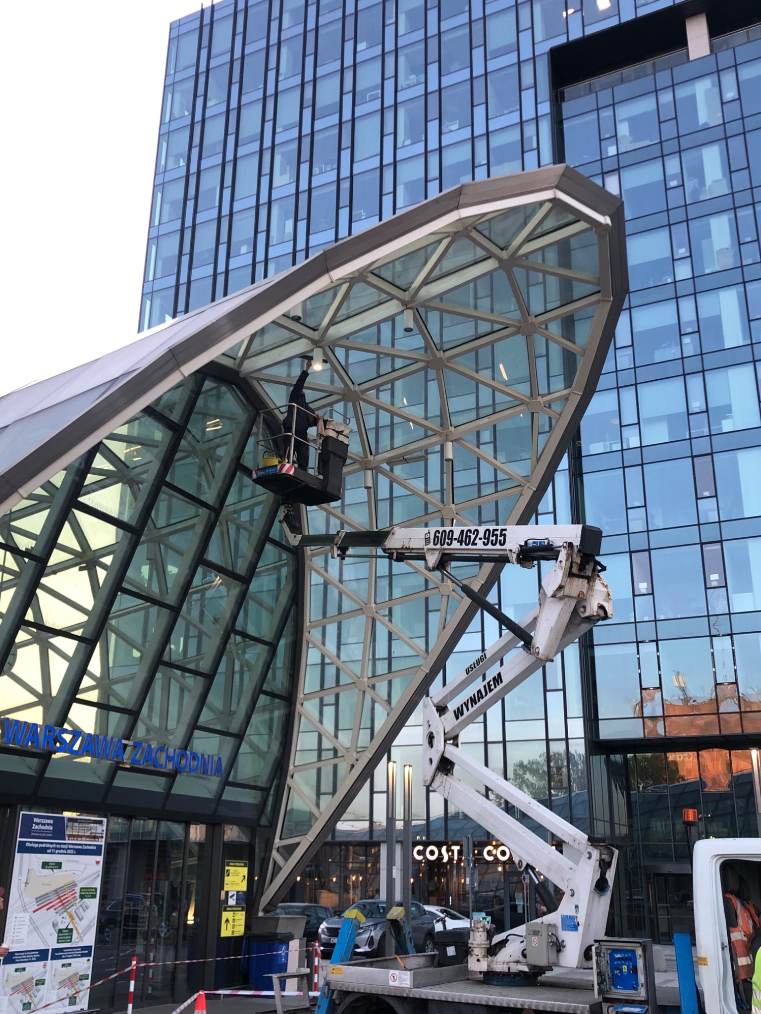 Pracownik w podnośniku czyści szklaną konstrukcję nad wejściem do budynku w Gostyninie. Widoczny numer telefonu na ramieniu podnośnika i fragment elewacji.