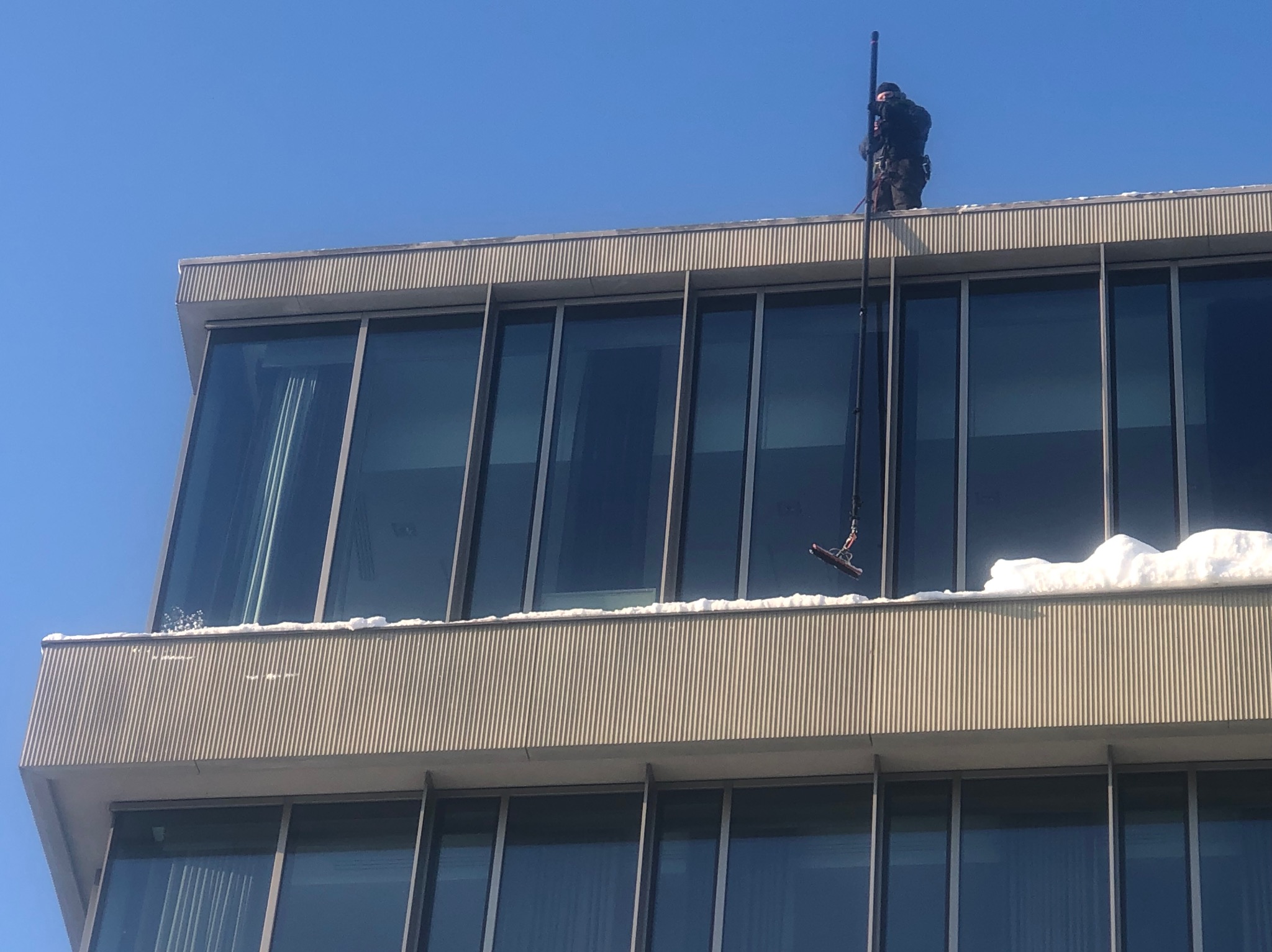 Pracownik w uprzęży na dachu budynku biurowego, używający długiej lancy do mycia okien. Widoczny śnieg na krawędzi dachu i błękitne niebo w tle.