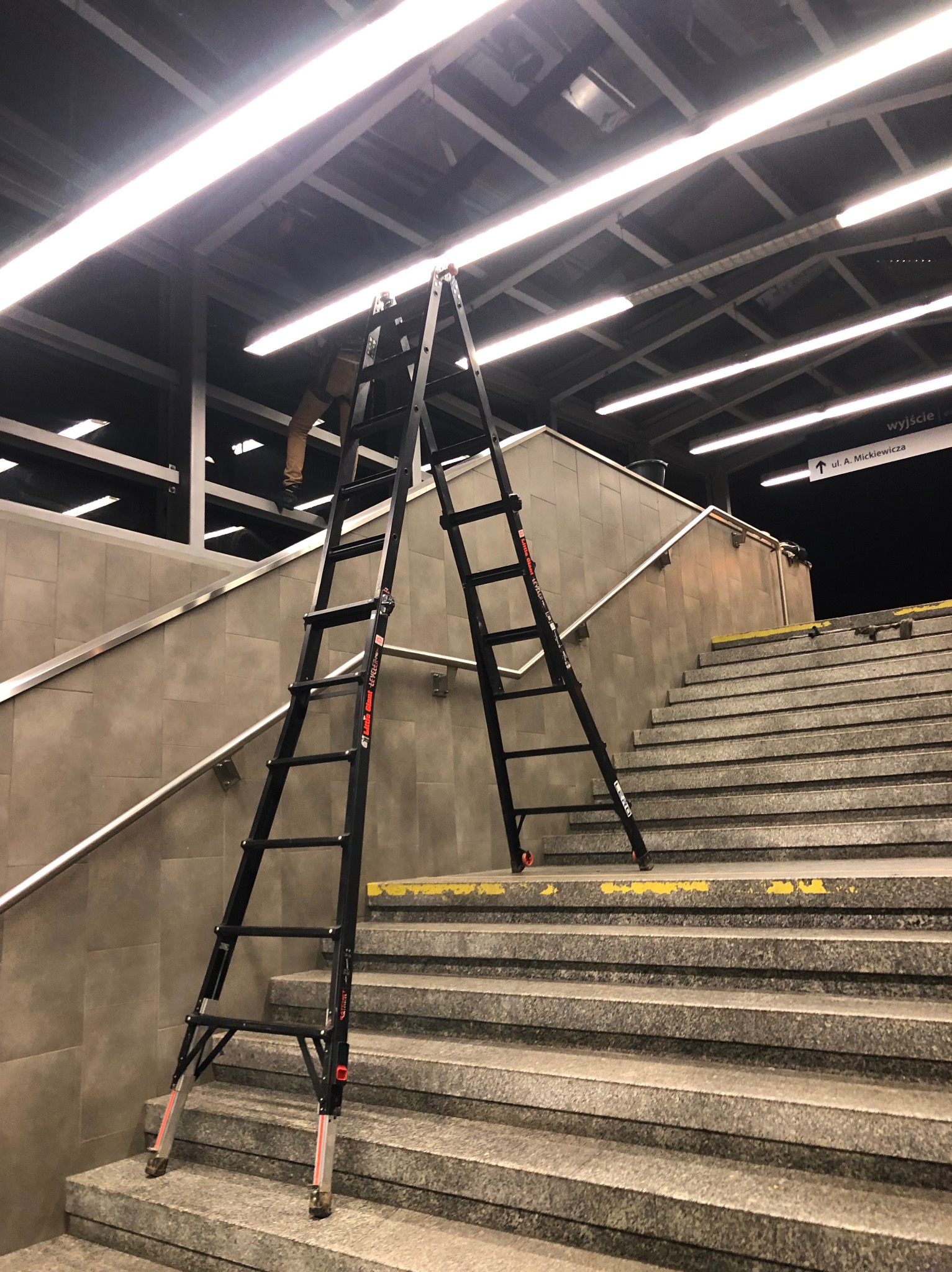 Rozstawiona czarna drabina na schodach wewnątrz budynku, prawdopodobnie w trakcie prac konserwacyjnych oświetlenia sufitowego. Widoczne nogi osoby na drabinie.