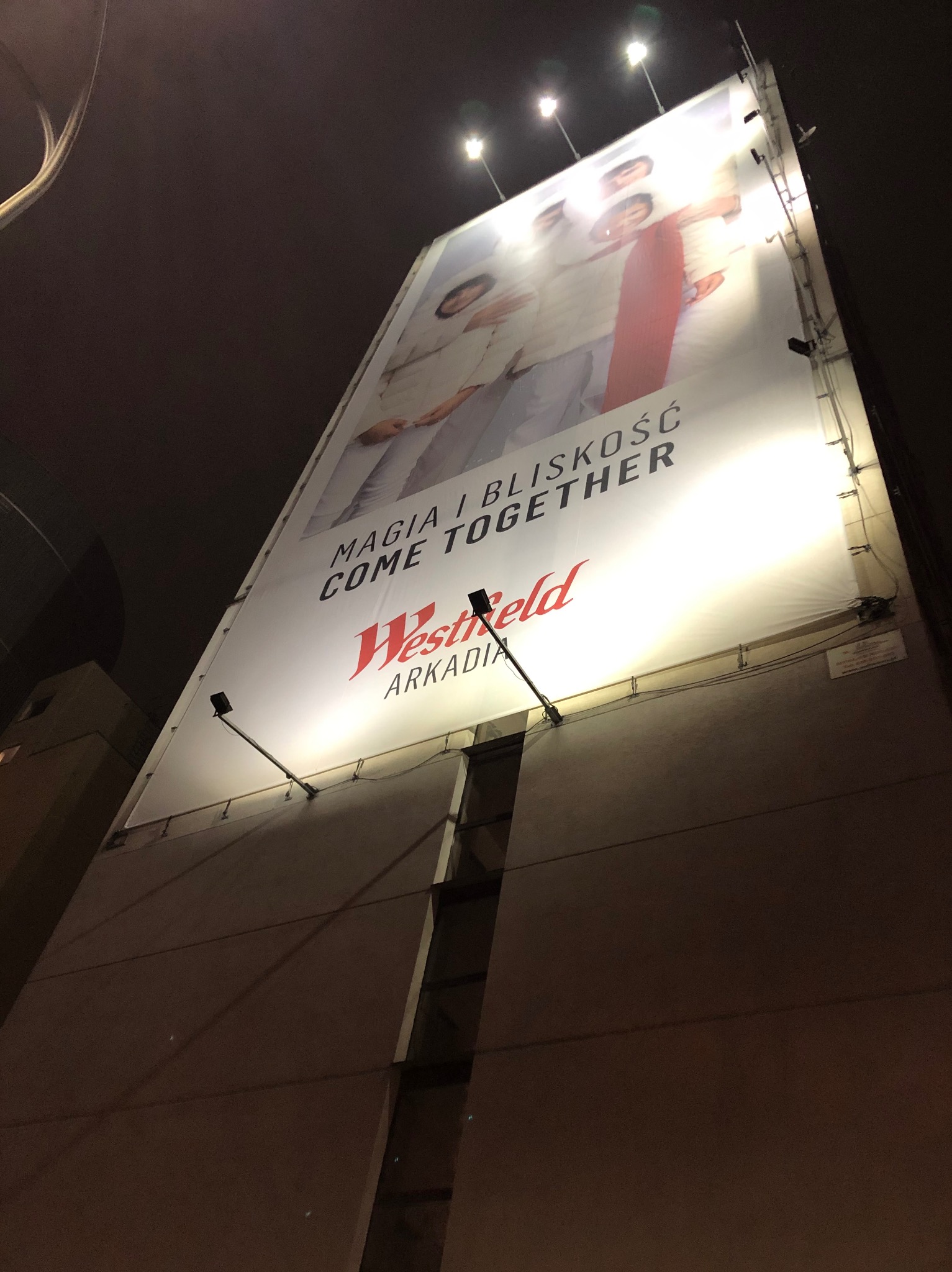 Wysoki billboard reklamowy Westfield Arkadia oświetlony nocą, z hasłem 'Magia i bliskość. Come together' i wizerunkiem ludzi. Ujęcie z dołu, widoczne mocowania i oświetlenie.