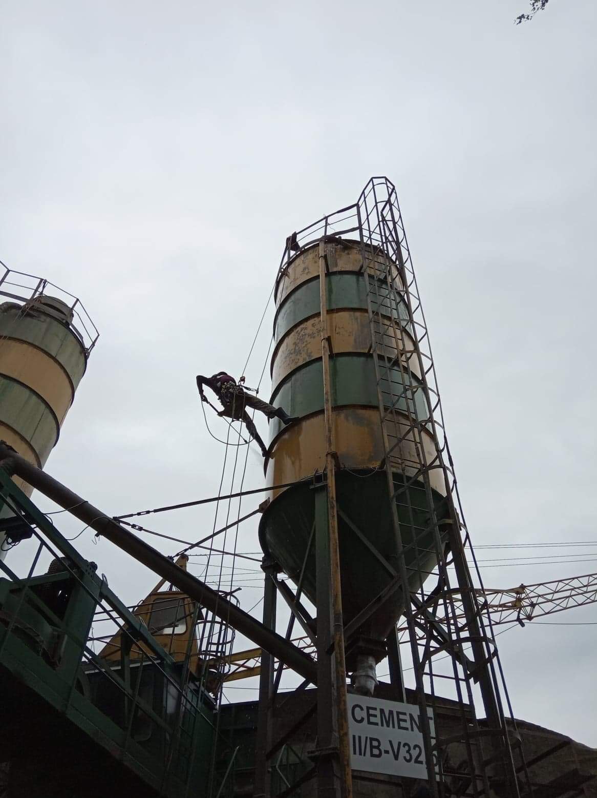 Pracownik na linie, zabezpieczony uprzężą, wykonuje prace na wysokości na silosie cementowym. Widoczne drabiny, liny i konstrukcja stalowa. Pochmurne niebo w tle.