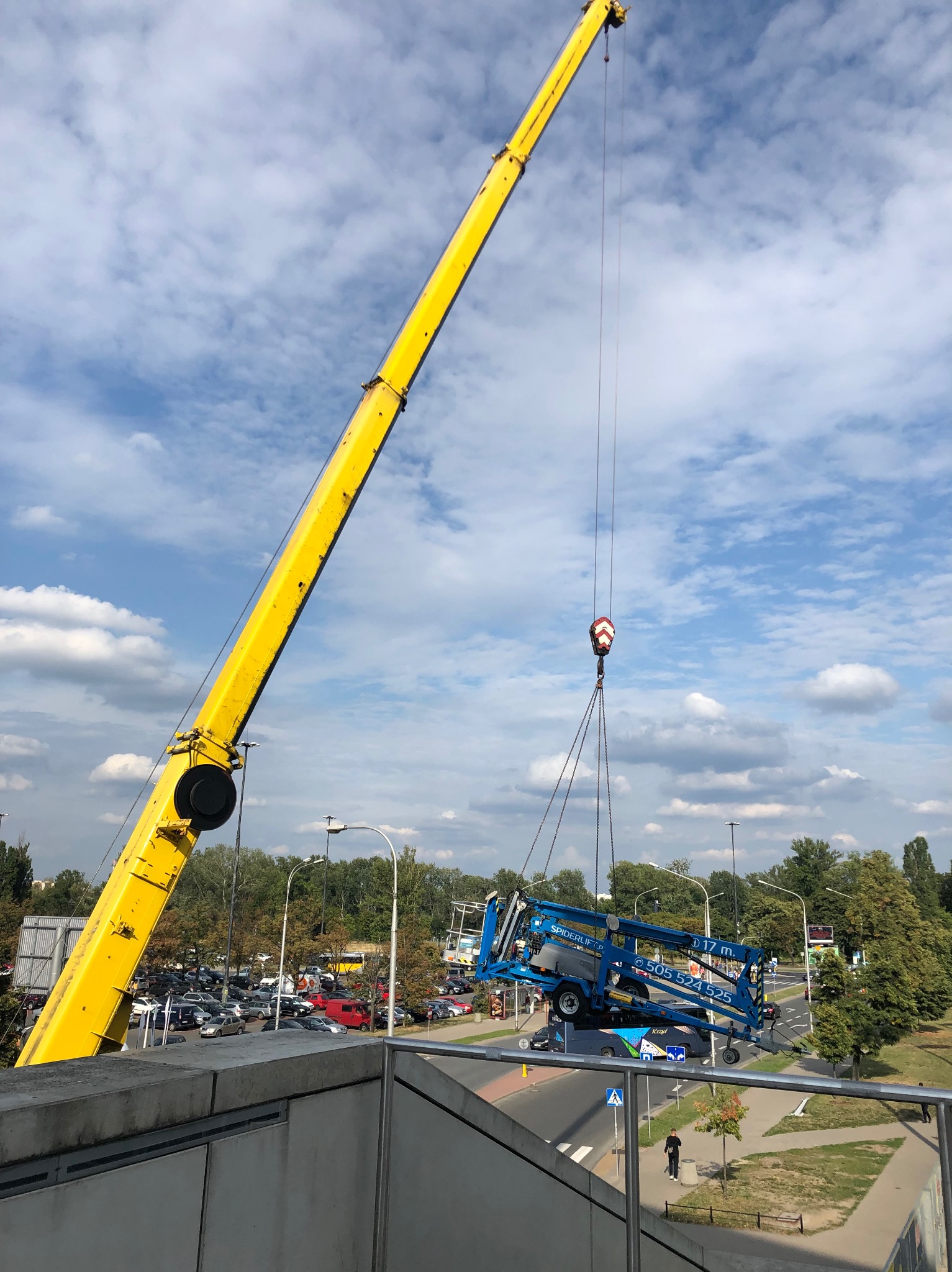 Żółty dźwig podnosi niebieski podnośnik koszowy nad ulicą. Widok z góry, błękitne niebo z chmurami w tle. Transport podnośnika.