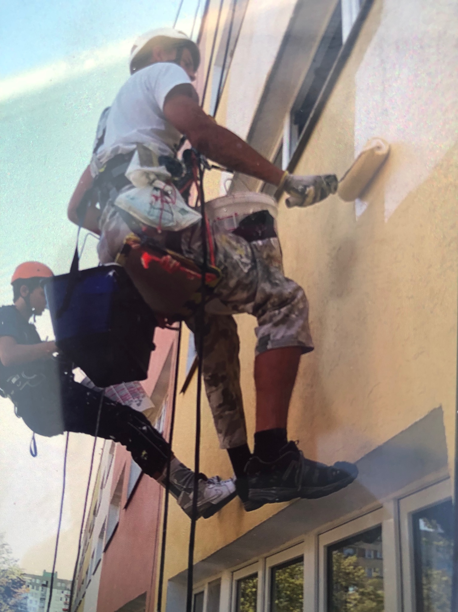 Dwóch pracowników na linach maluje elewację budynku wałkiem. Jeden maluje, drugi asekuruje. Widoczne okna i fragmenty innych budynków w tle.