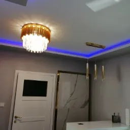 Nowoczesne oświetlenie wnętrza: złoty żyrandol z kryształkami i trzy złote lampy wiszące, podświetlenie LED w kolorze niebieskim, białe drzwi z czarnym szkłem i marmurowa ściana.