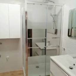 Nowoczesna łazienka z kabiną prysznicową z częściowo matowego szkła, białą szafką wiszącą i umywalką z szafką, wykończona drewnopodobną podłogą, w odbiciu w szybie widoczna kolejna umywalka.