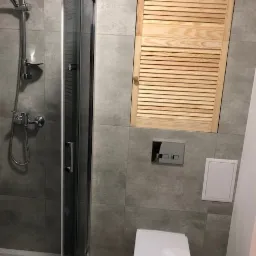 Nowoczesna łazienka z kabiną prysznicową ze szklanymi drzwiami, toaletą wiszącą i drewnianą szafką z żaluzjowymi drzwiczkami w górnej części ściany wyłożonej szarymi płytkami.