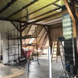 Poddasze w trakcie prac wykończeniowych: widoczne ocieplenie wełną mineralną, rusztowanie, ceglany komin, drewniane belki konstrukcyjne oraz częściowo ułożona podłoga z płytek.