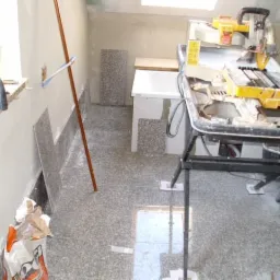 Ułożona podłoga z szarych płytek imitujących kamień, fragment ściany z przyklejonymi płytkami, elektryczna przecinarka do płytek i paczka kleju w rogu pomieszczenia w trakcie prac wykończeniowych.