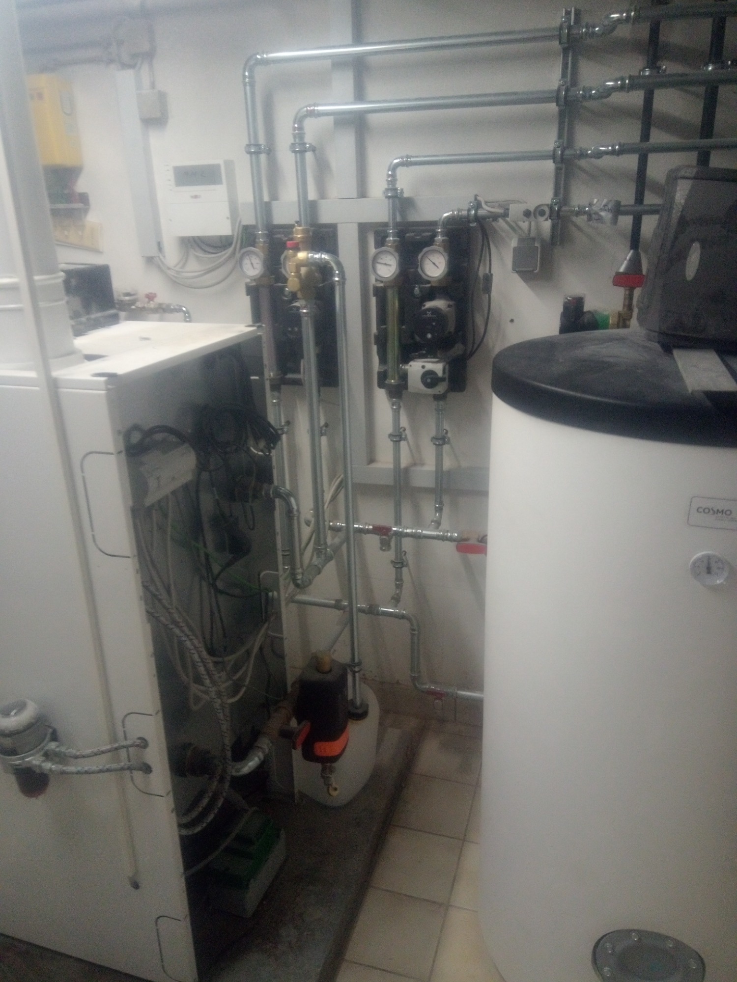 Widok na kotłownię z białym zasobnikiem ciepłej wody marki Cosmo, licznymi rurami, manometrami i osprzętem, ukazujący skomplikowaną instalację grzewczą w pomieszczeniu technicznym.