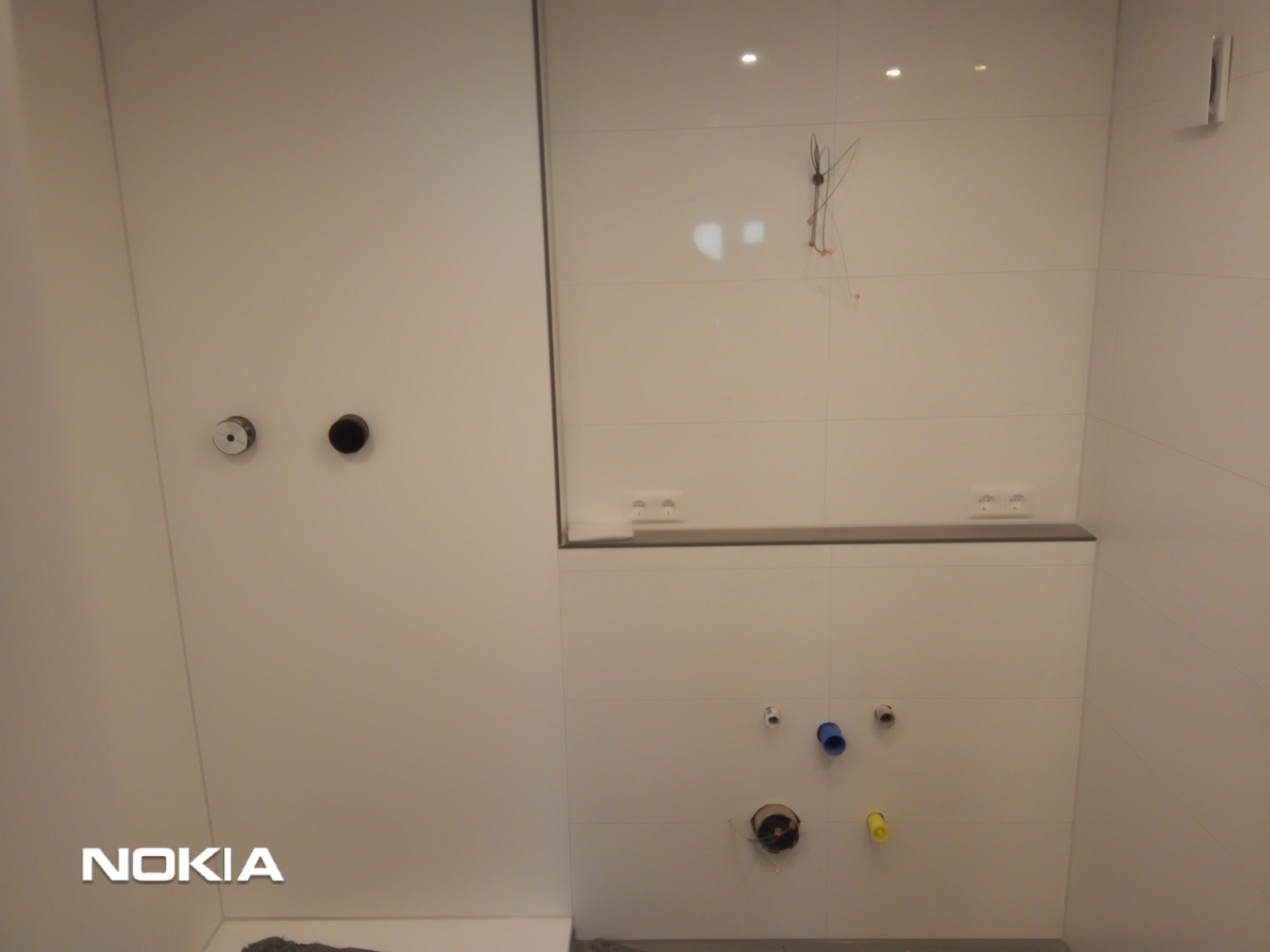 Świeżo wykafelkowana, minimalistyczna łazienka z przygotowanymi punktami instalacyjnymi: gniazdka elektryczne nad szarym blatem, wyprowadzenia rur z niebieskim i żółtym zaślepieniem oraz otwór...