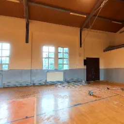 Sala gimnastyczna w trakcie odświeżania: pomalowane ściany na kremowo powyżej szarego pasa, drewniany parkiet, rusztowanie przy oknie, widoczny sprzęt do sprzątania i worki z materiałami...